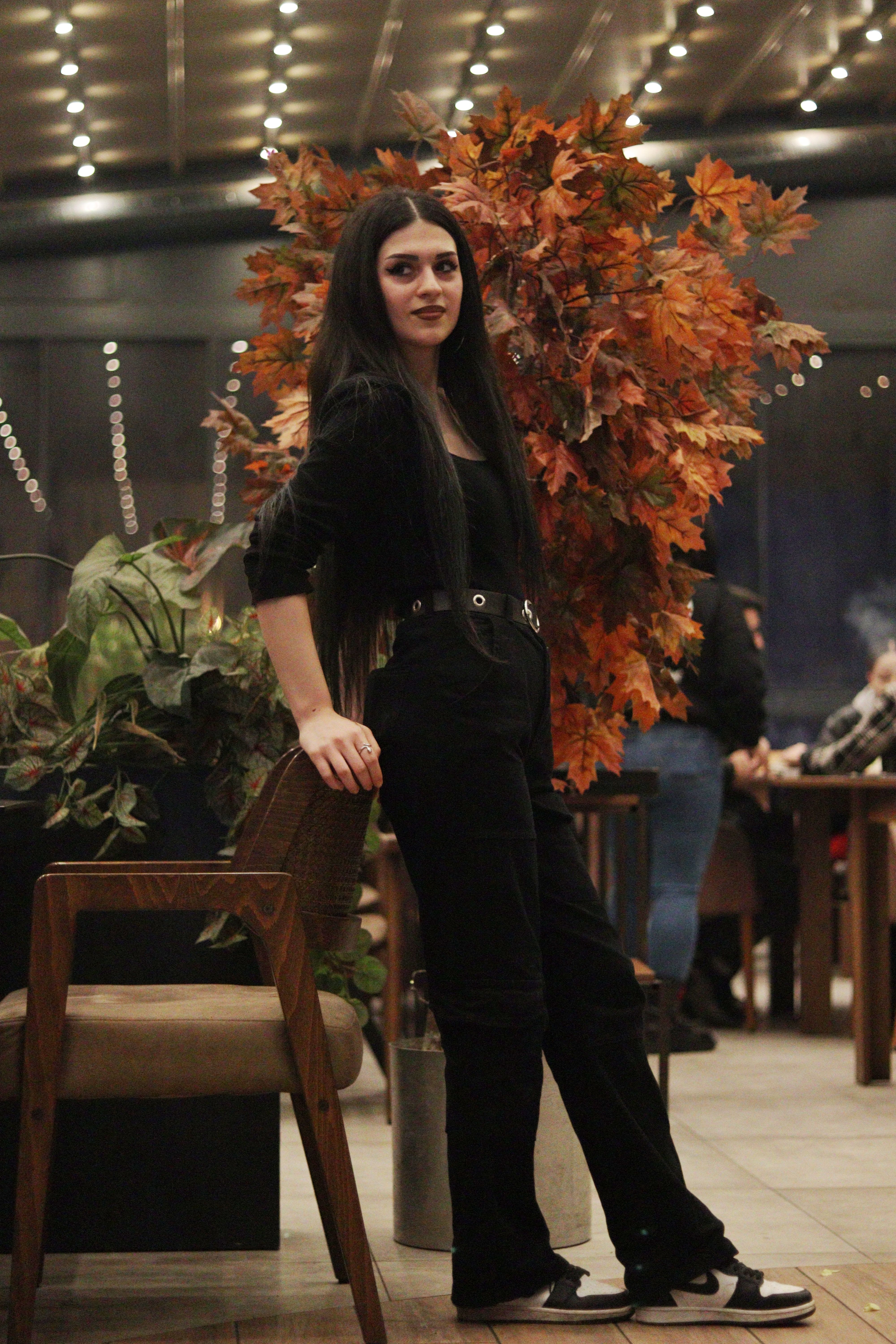 Woman posing indoors beside autumn leaves decor in a cozy venue.