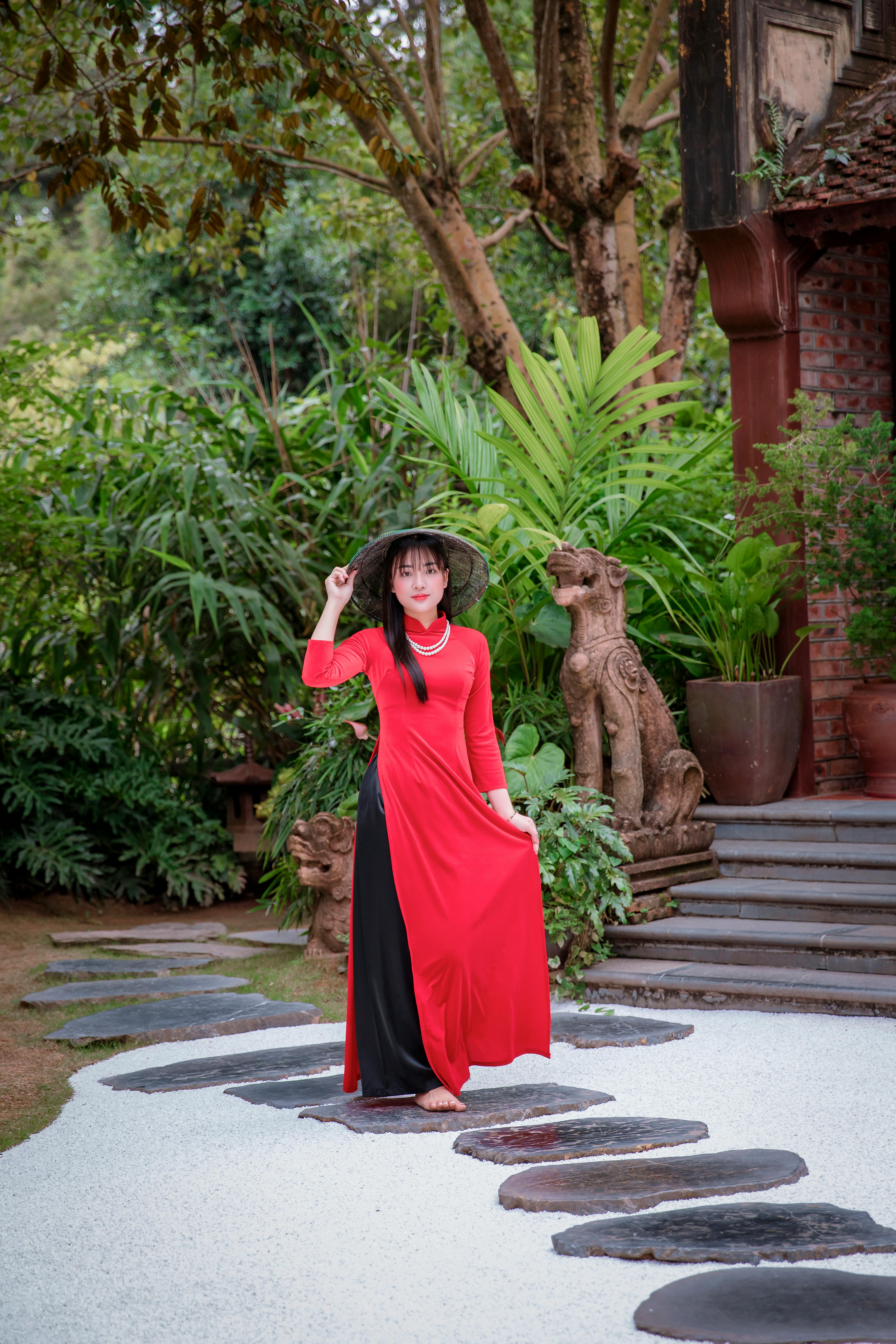 A woman poses in a vibrant red dress outdoors, surrounded by lush greenery and stone sculptures.