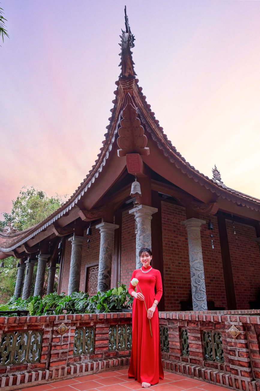 A woman in a red Ao Dai poses at a Vietnamese temple during sunset, showcasing cultural beauty.