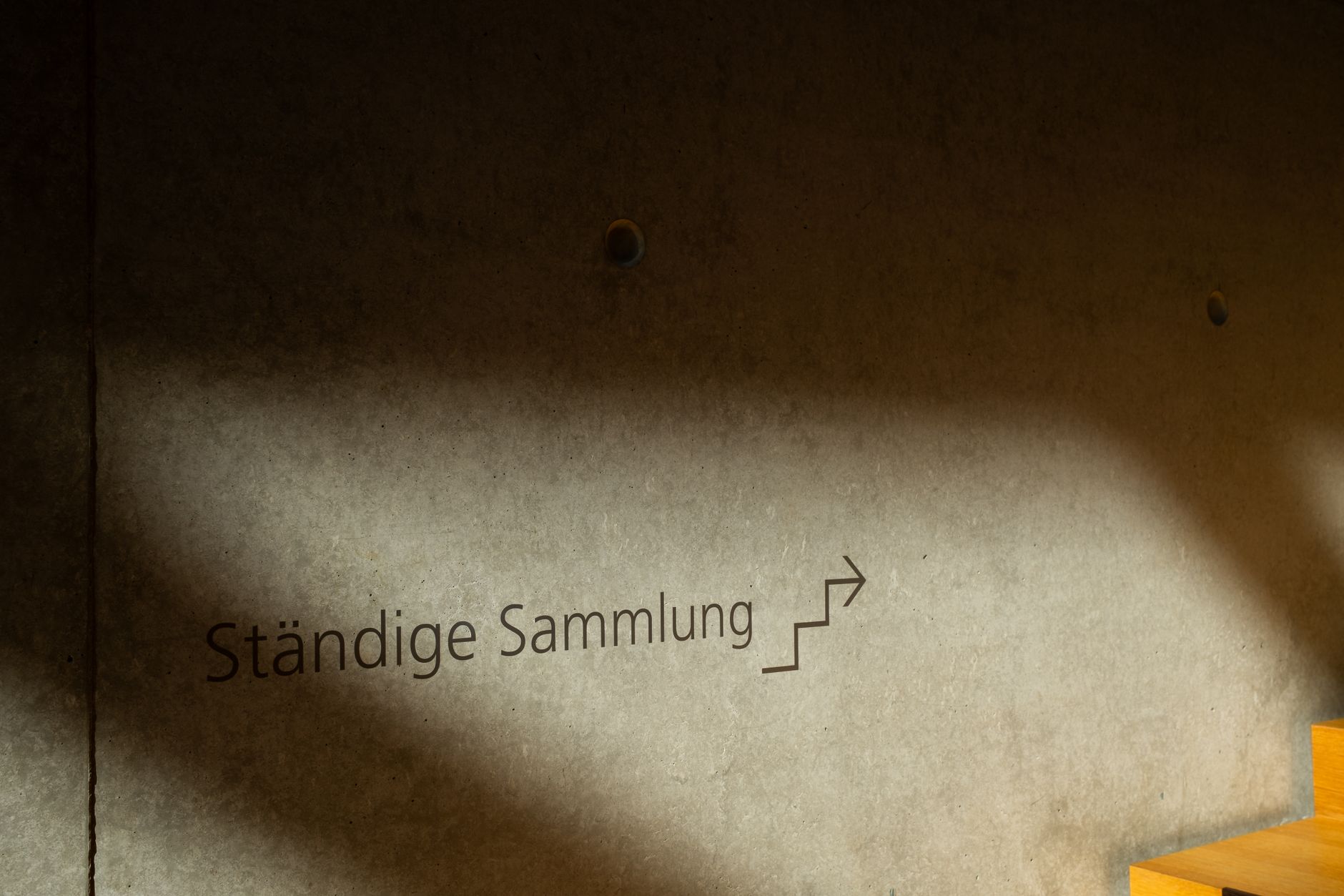 Concrete wall featuring German text 'Ständige Sammlung' illuminated by sunlight, with a staircase.