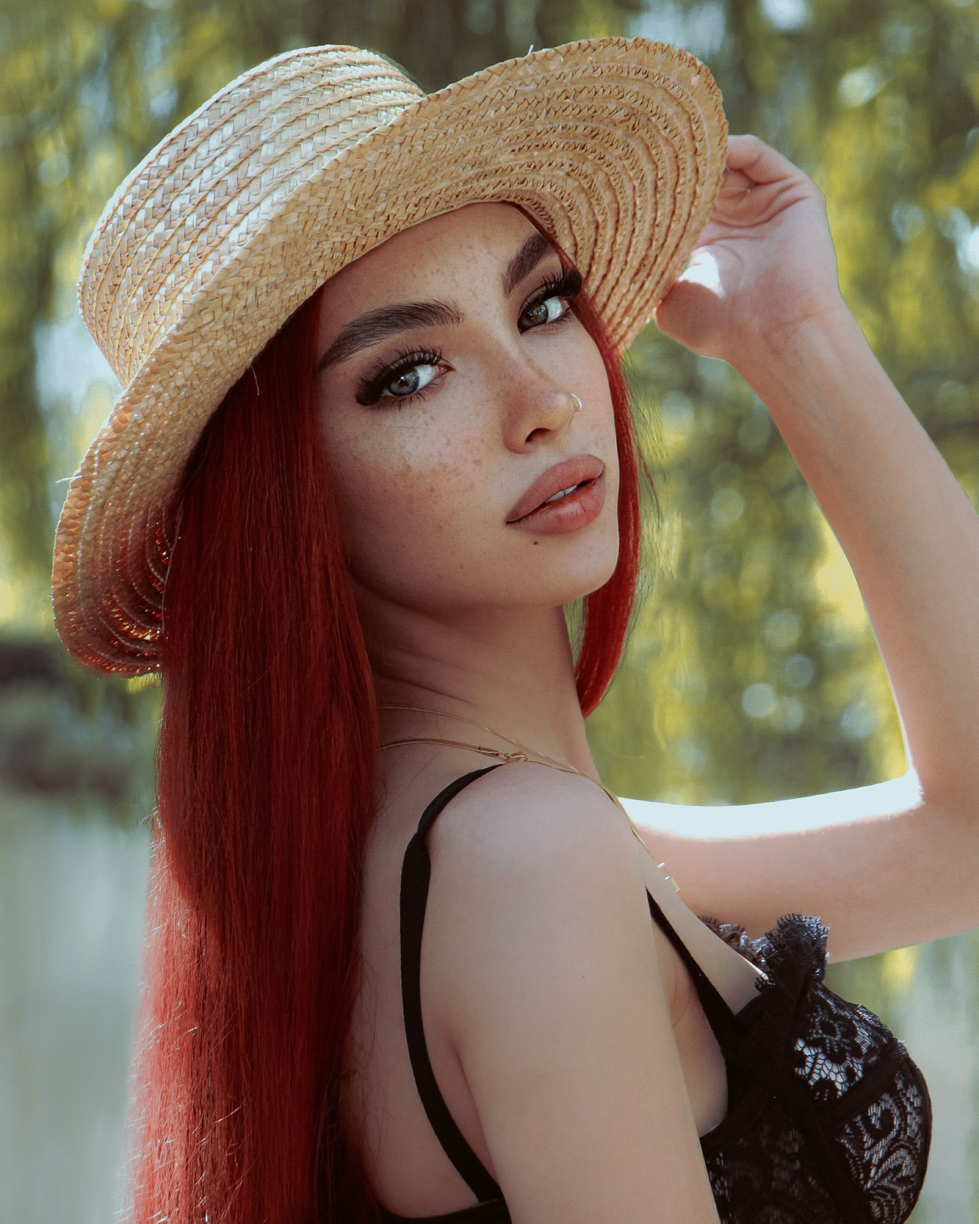 Fashionable woman with red hair poses outdoors in summer sunlight wearing a straw hat.
