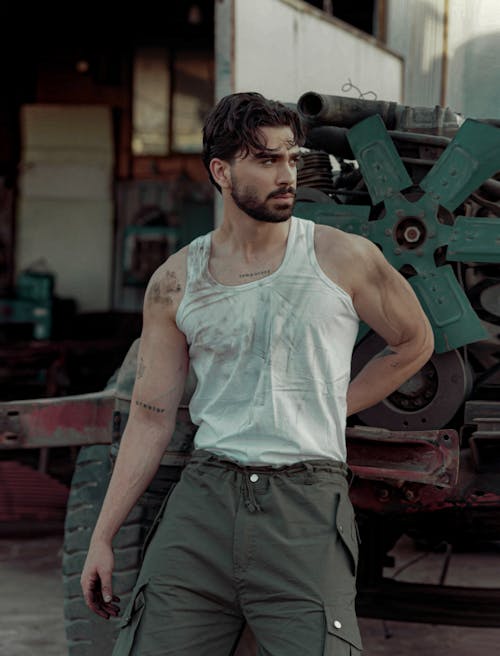 Man in a Dirty Tank Top and Green Cargo Pants in Front of an Old Truck Engine