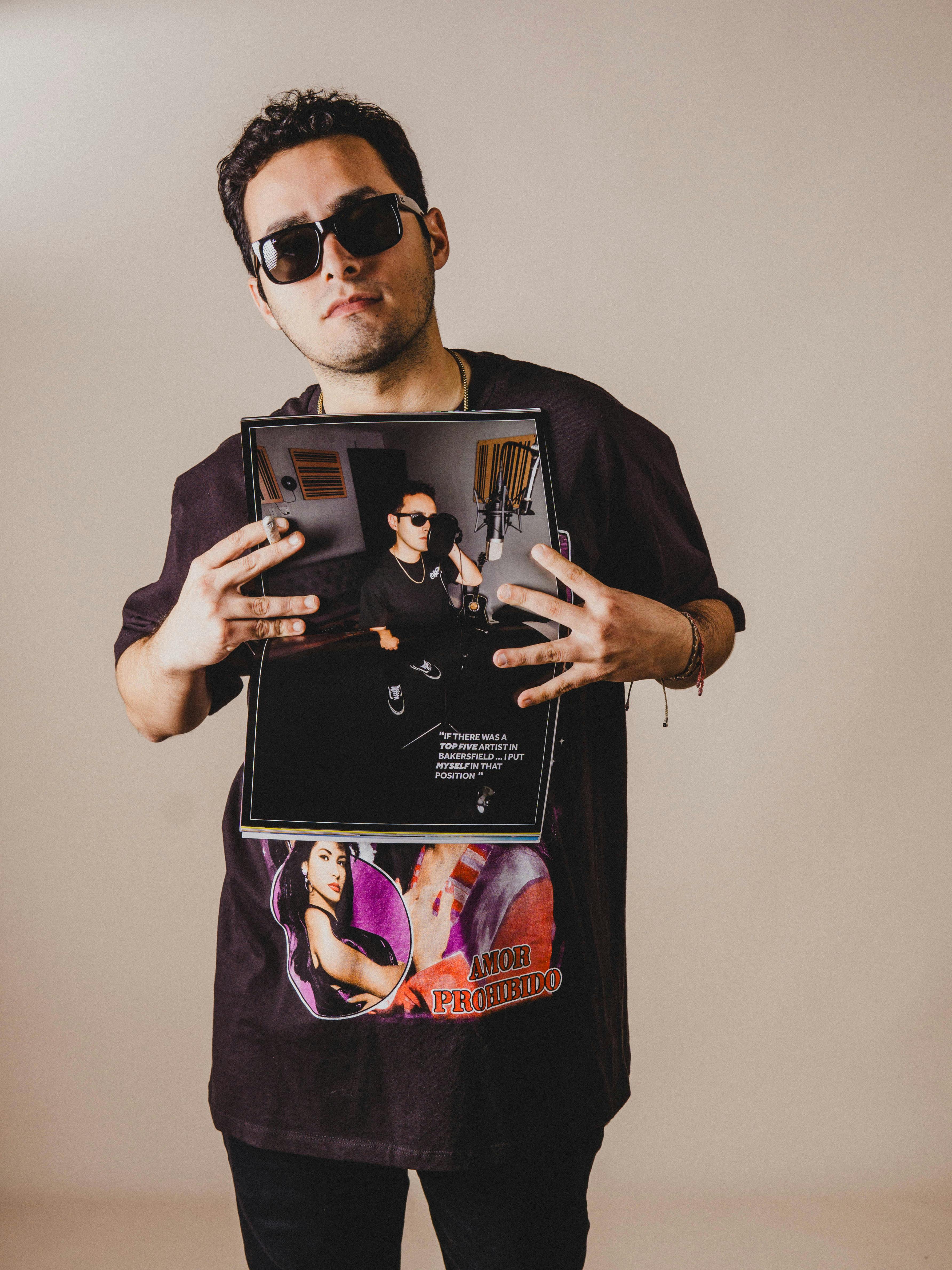 Young man in sunglasses holding a magazine, wearing a graphic t-shirt in a studio.