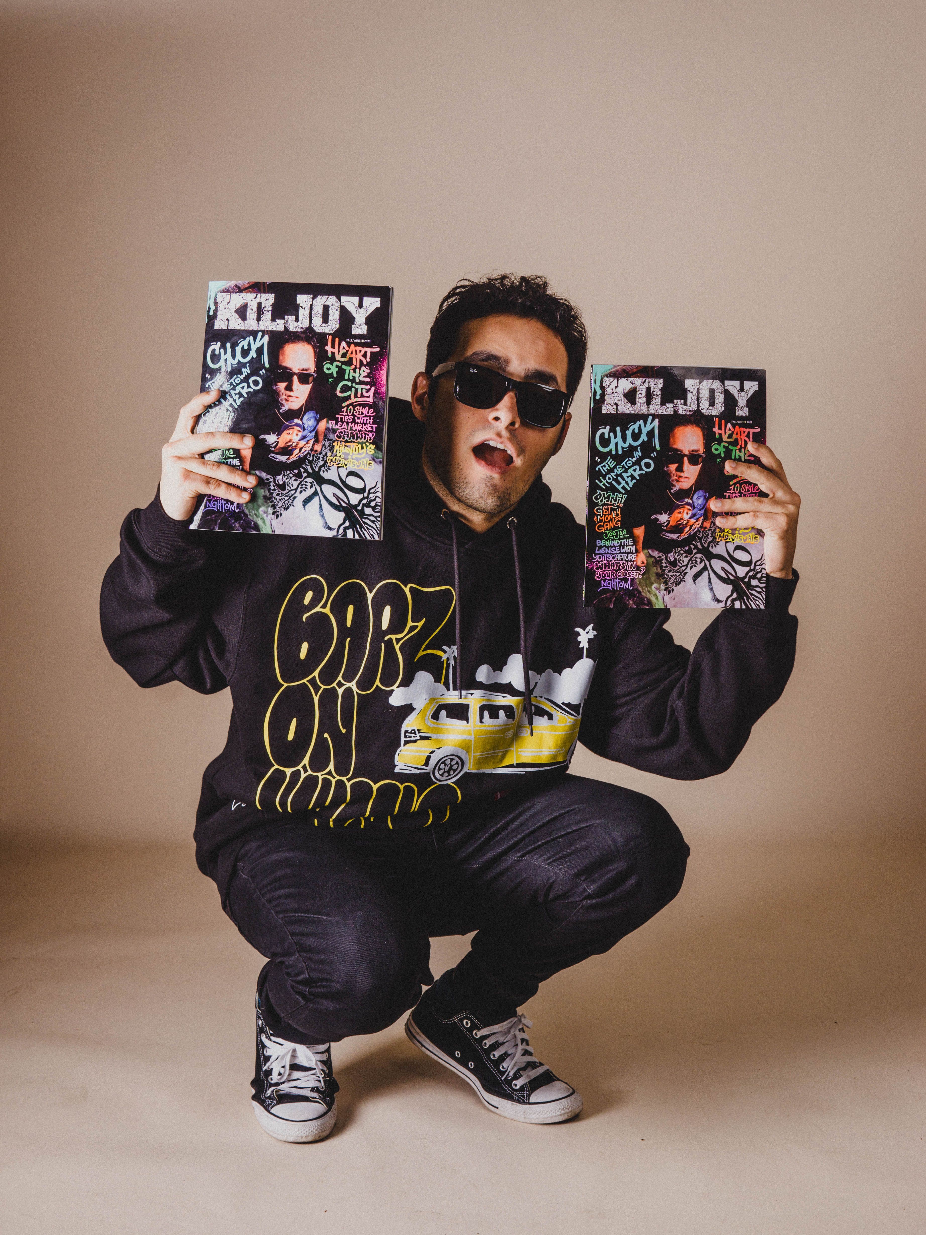 Young man in streetwear holding Kiljoy magazines in a stylish studio photo.
