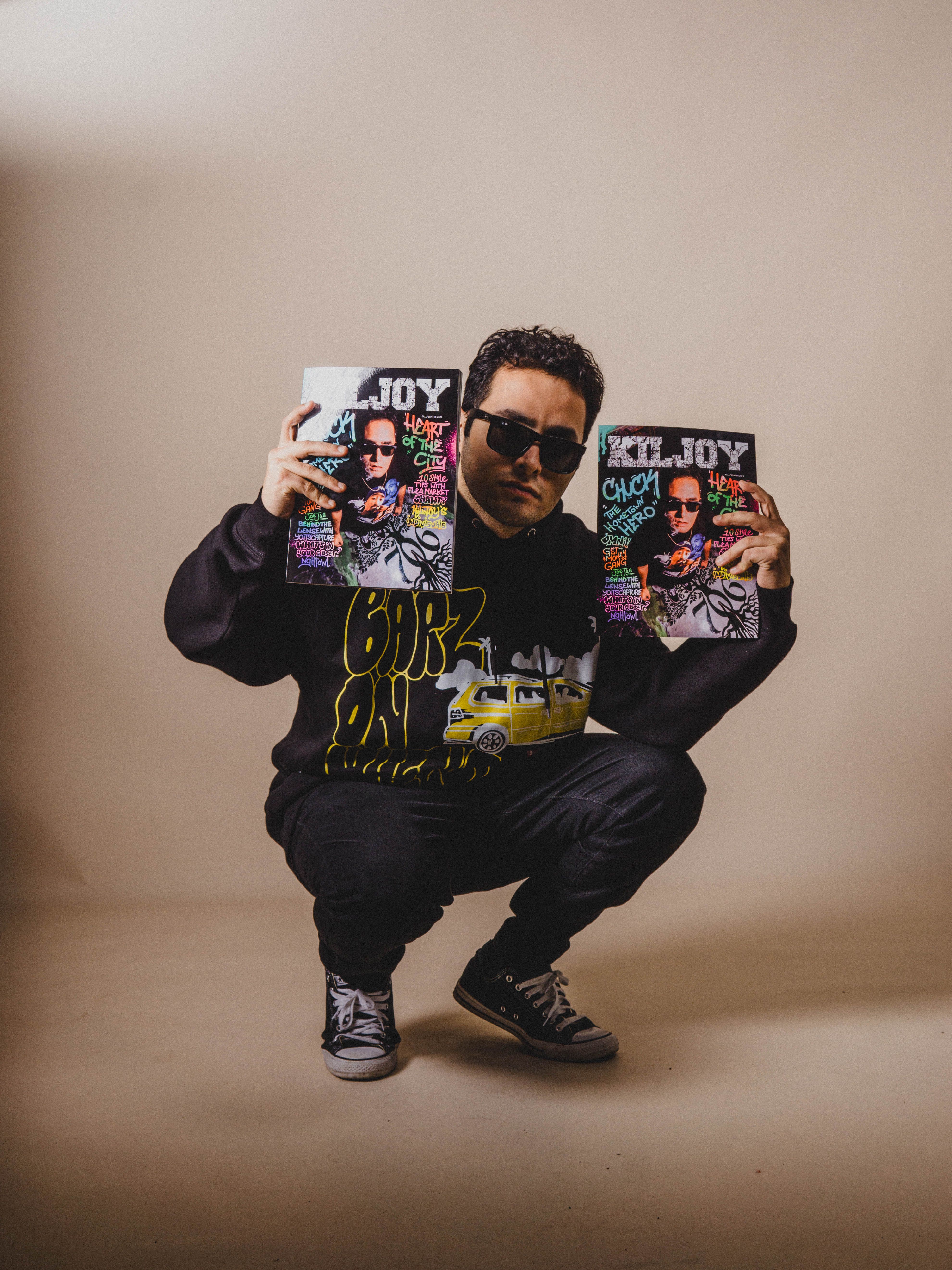 Stylish man crouching in a studio holding vibrant graphic magazines, wearing a hoodie and sunglasses.
