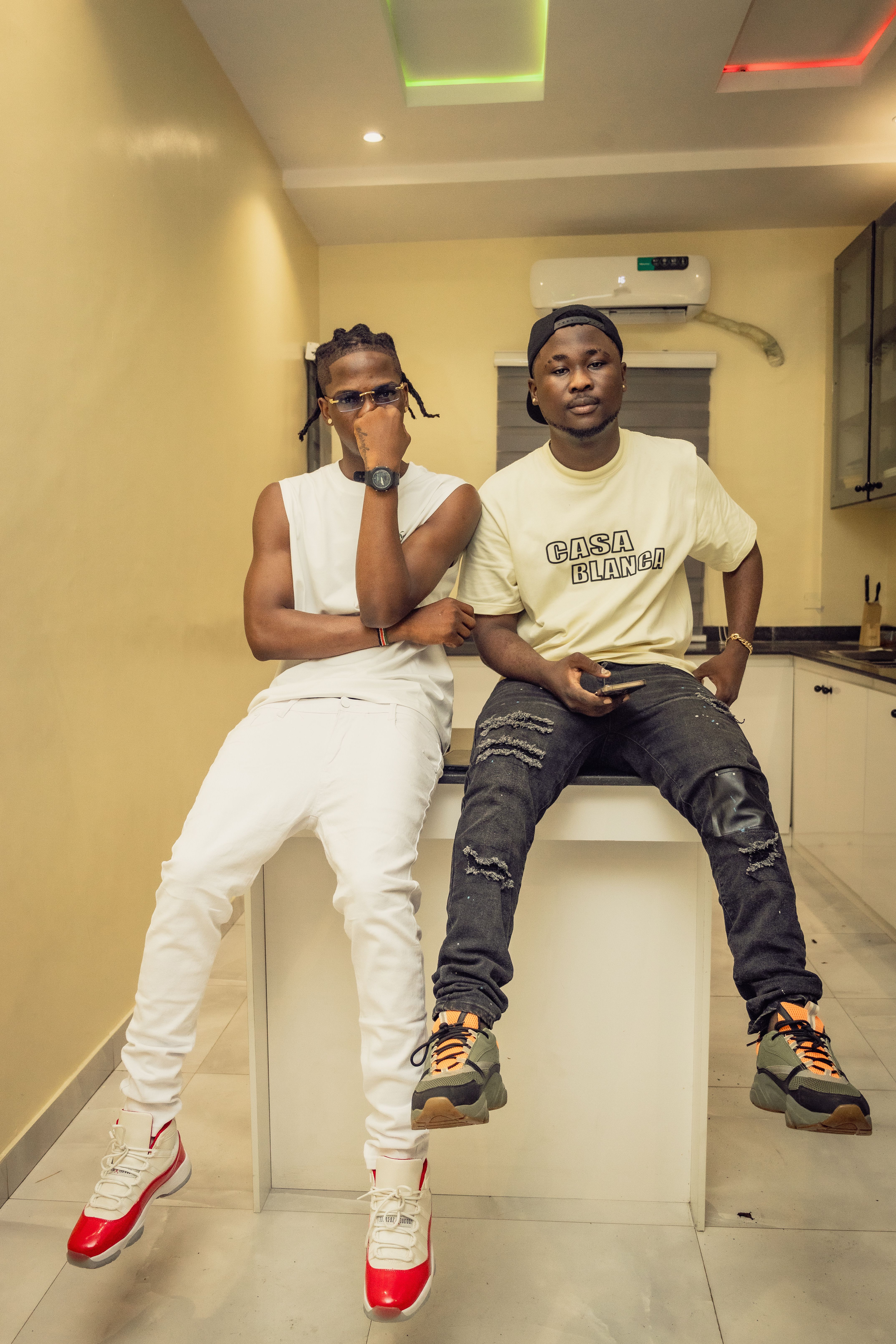 Two young men in streetwear pose stylishly atop a kitchen countertop.