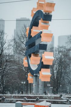 Modern Euro sculpture in a snowy town square with urban skyscrapers.