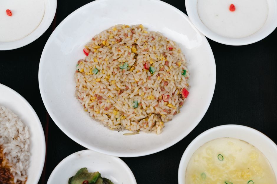 A wide steel griddle covered in golden fried rice studded with scrambled egg, peas, carrots, and scallions, with a metal spatula pushing a crispy edge.