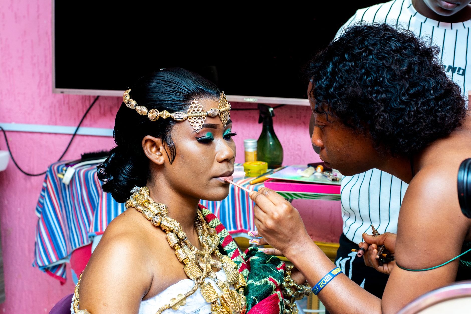 A bride in traditional attire getting her makeup done by a beautician in a colorful indoor setting.