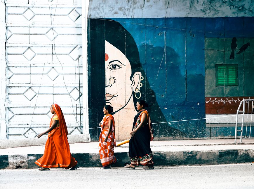 Three Indian women in traditional saris walk past a vibrant mural on a city sidewalk.