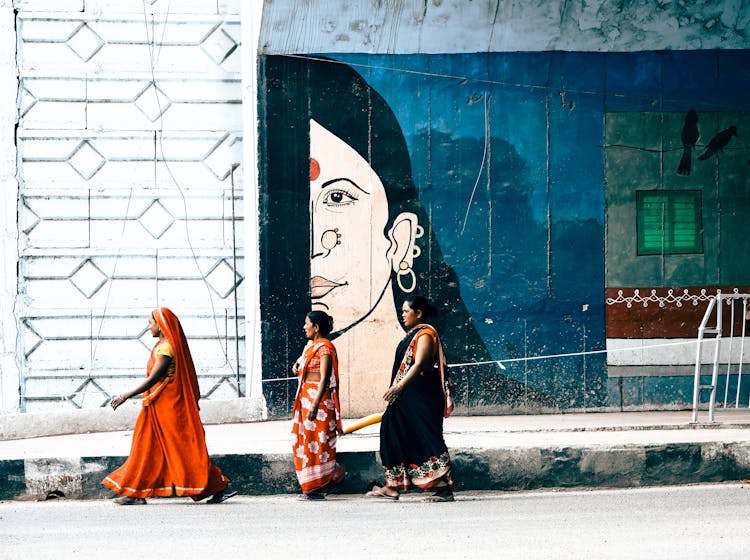 Women In Saris Walking On A Sidewalk Near A Building With A Mural 