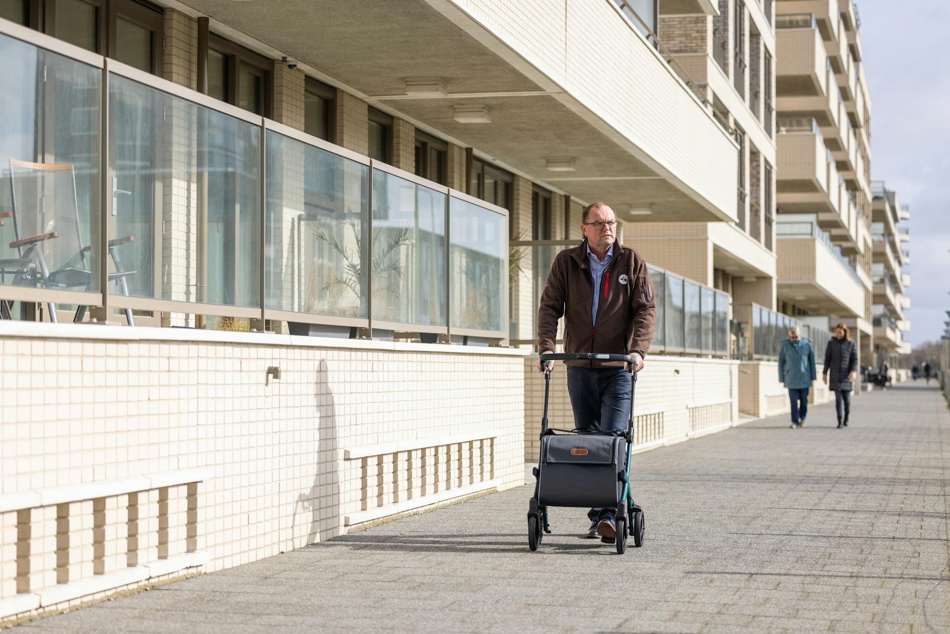 Senior man using rollator on a sunny city sidewalk beside residential buildings.