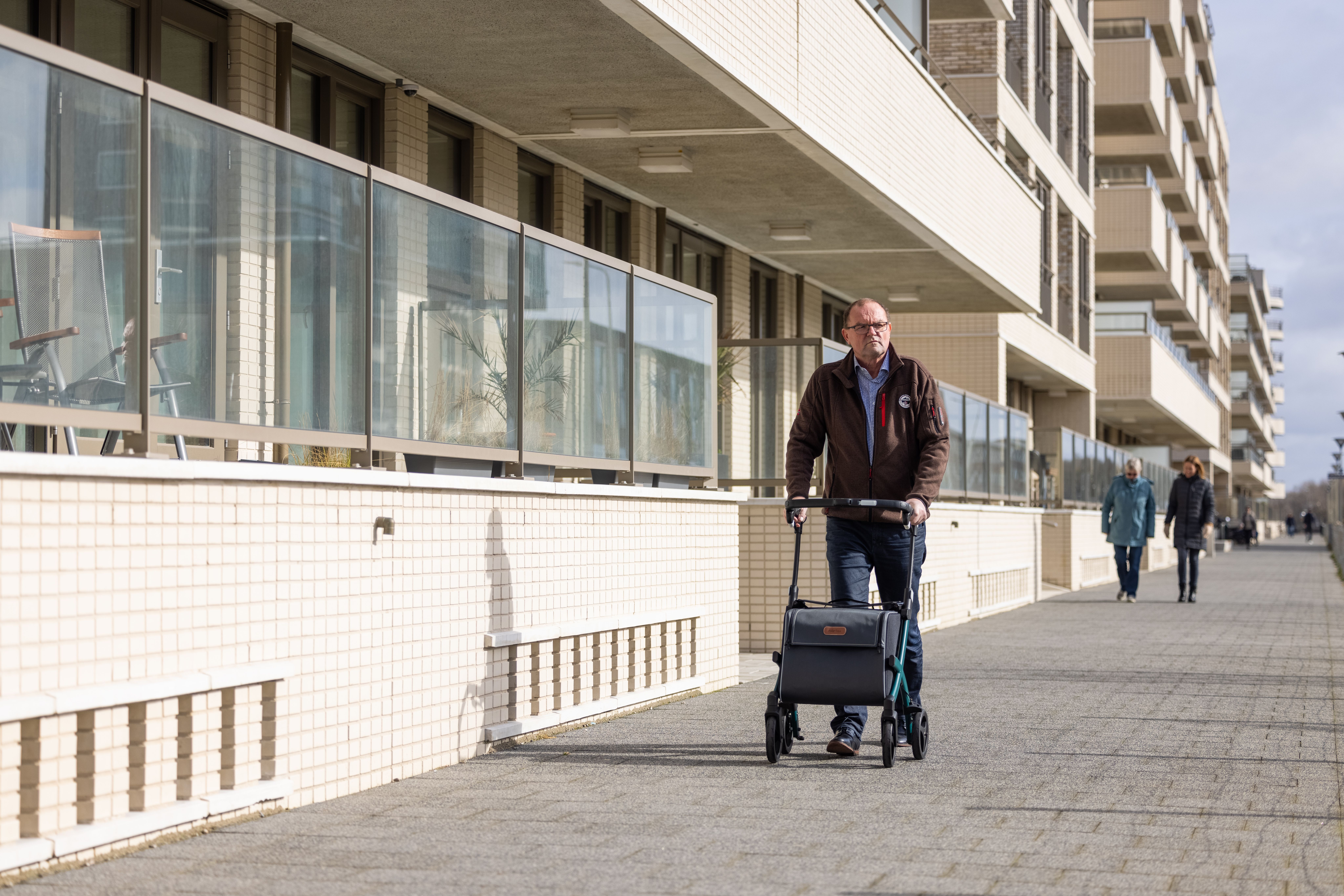 An elderly man using a rollator strolls down a city sidewalk near residential buildings.