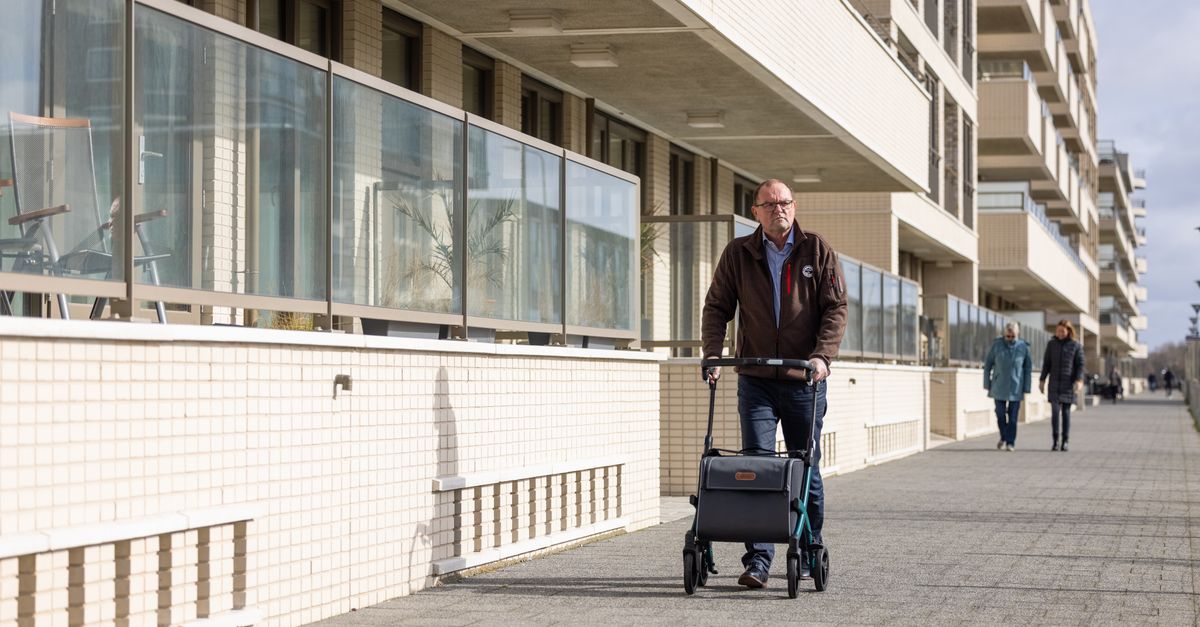 An elderly man using a rollator strolls down a city sidewalk near residential buildings.