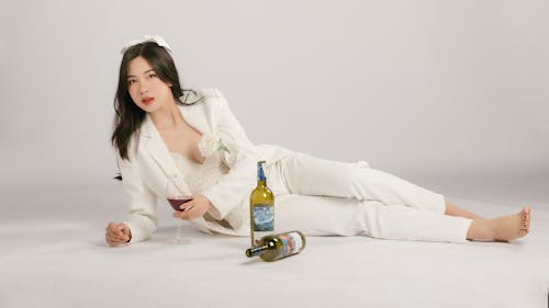 Young Woman in a White Outfit Posing on the Floor with Bottles of Wine 