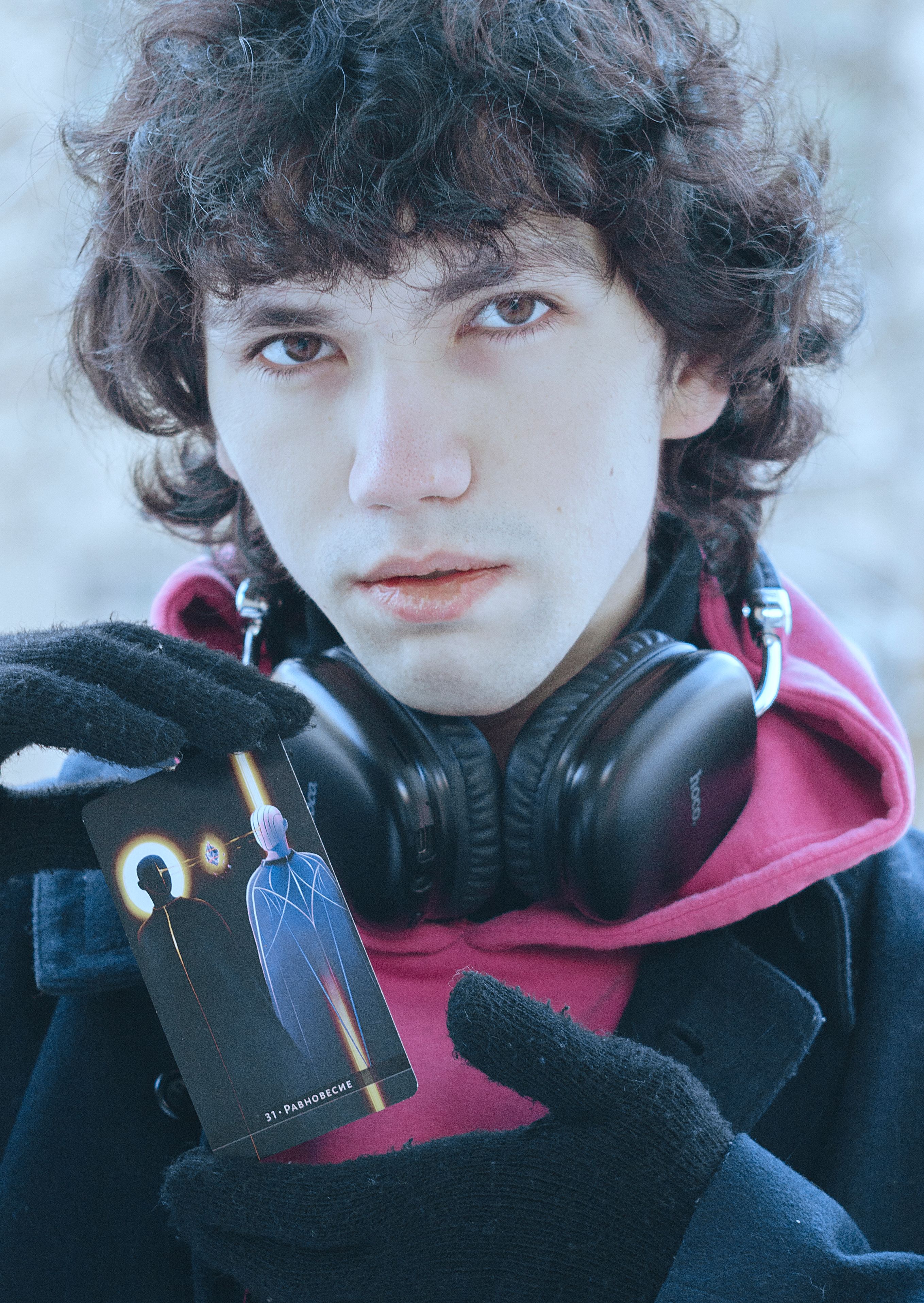 Portrait of a young man wearing headphones and gloves, holding a tarot card outdoors in winter.