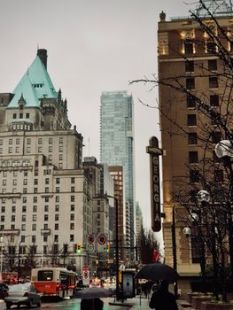 A rainy urban street scene in downtown Vancouver featuring notable skyscrapers and historic hotels.