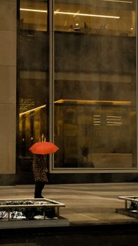 A woman stands with a red umbrella in front of a building on a rainy city street.