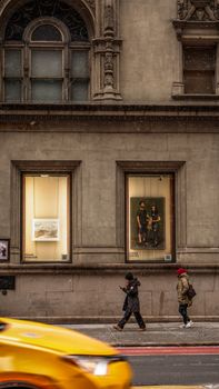 A winter street scene capturing people walking past a historic urban building with a yellow taxi blur.