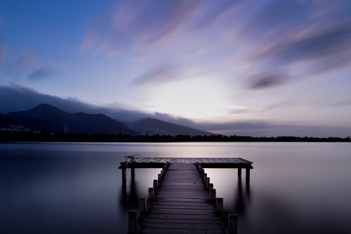 Free A tranquil view of a wooden pier extending into a calm lake during twilight with mountains in the background. Stock Photo
