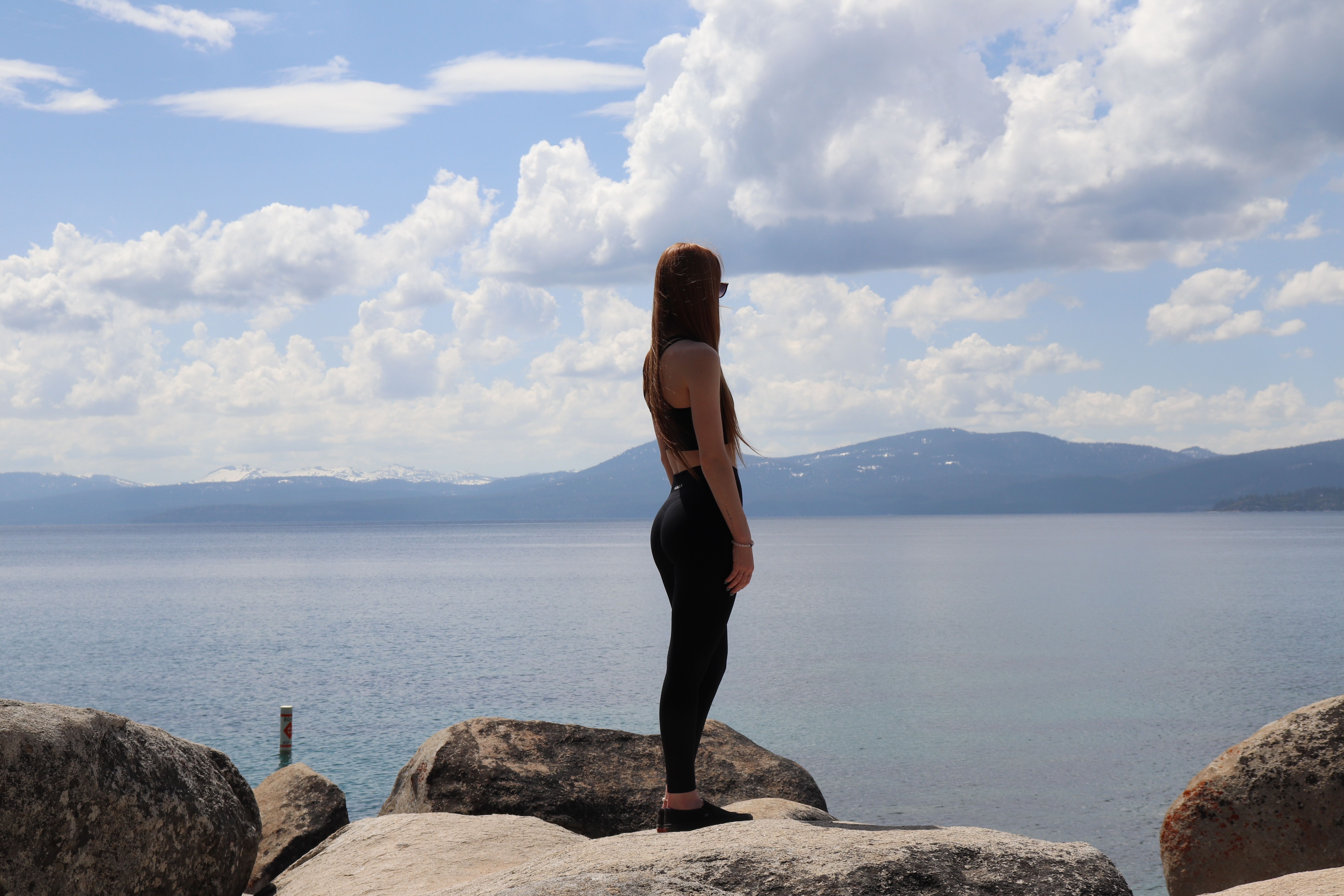 A woman in sportswear stands on rocks by a calm lake under a partly cloudy sky.