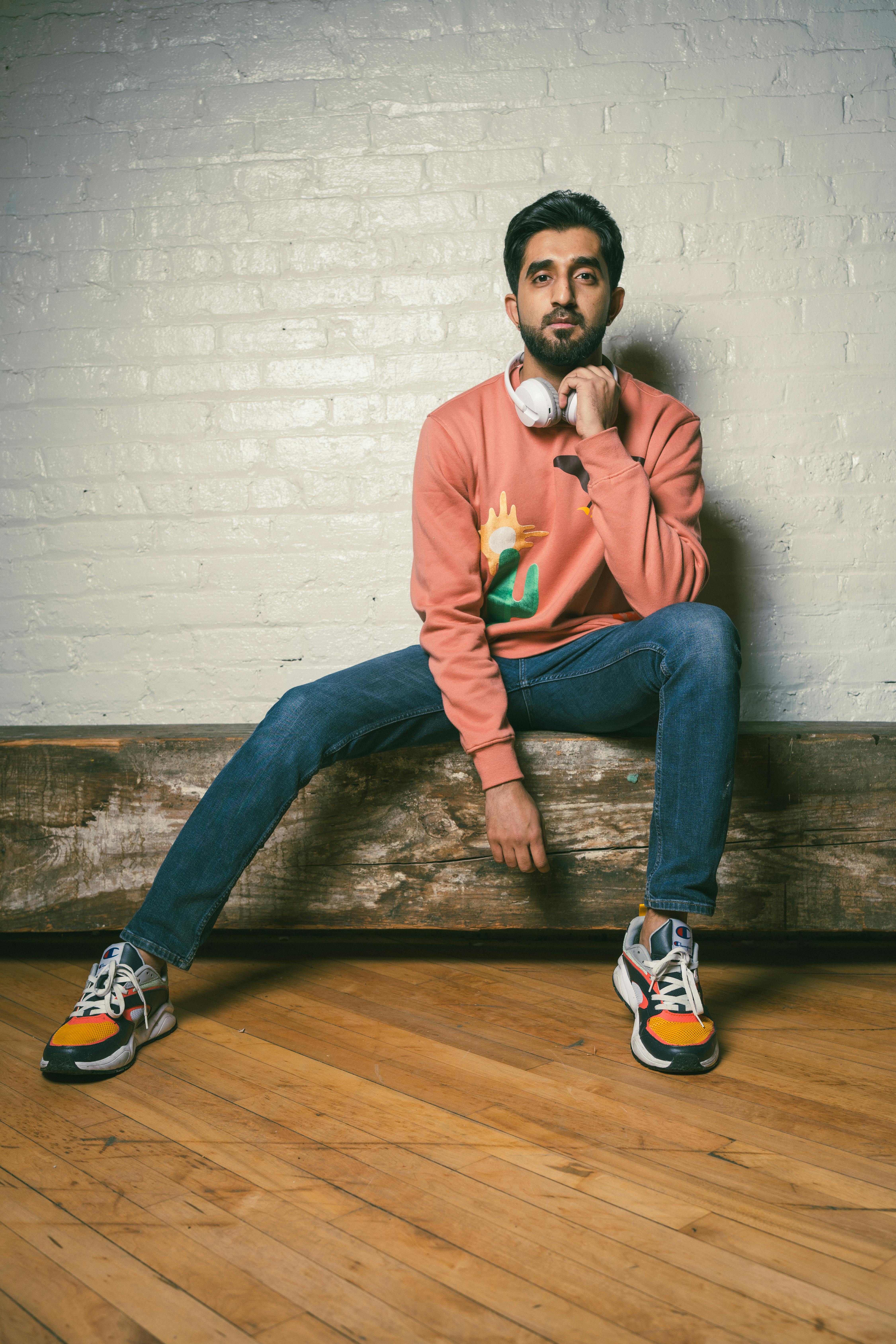 Man sitting against brick wall, wearing casual fashion with headphones and jeans.
