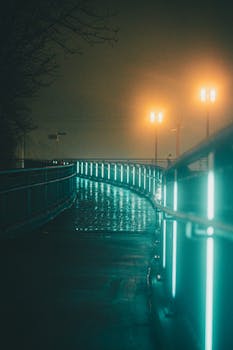 A moody night scene of an illuminated urban bridge shrouded in fog with glowing streetlamps and reflections.