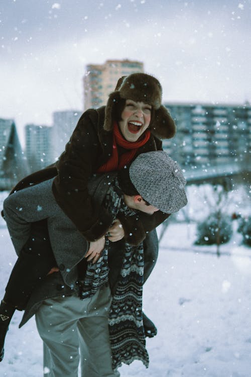 Laughing Woman in Fur Hat Sitting on Mans Back under Snowfall