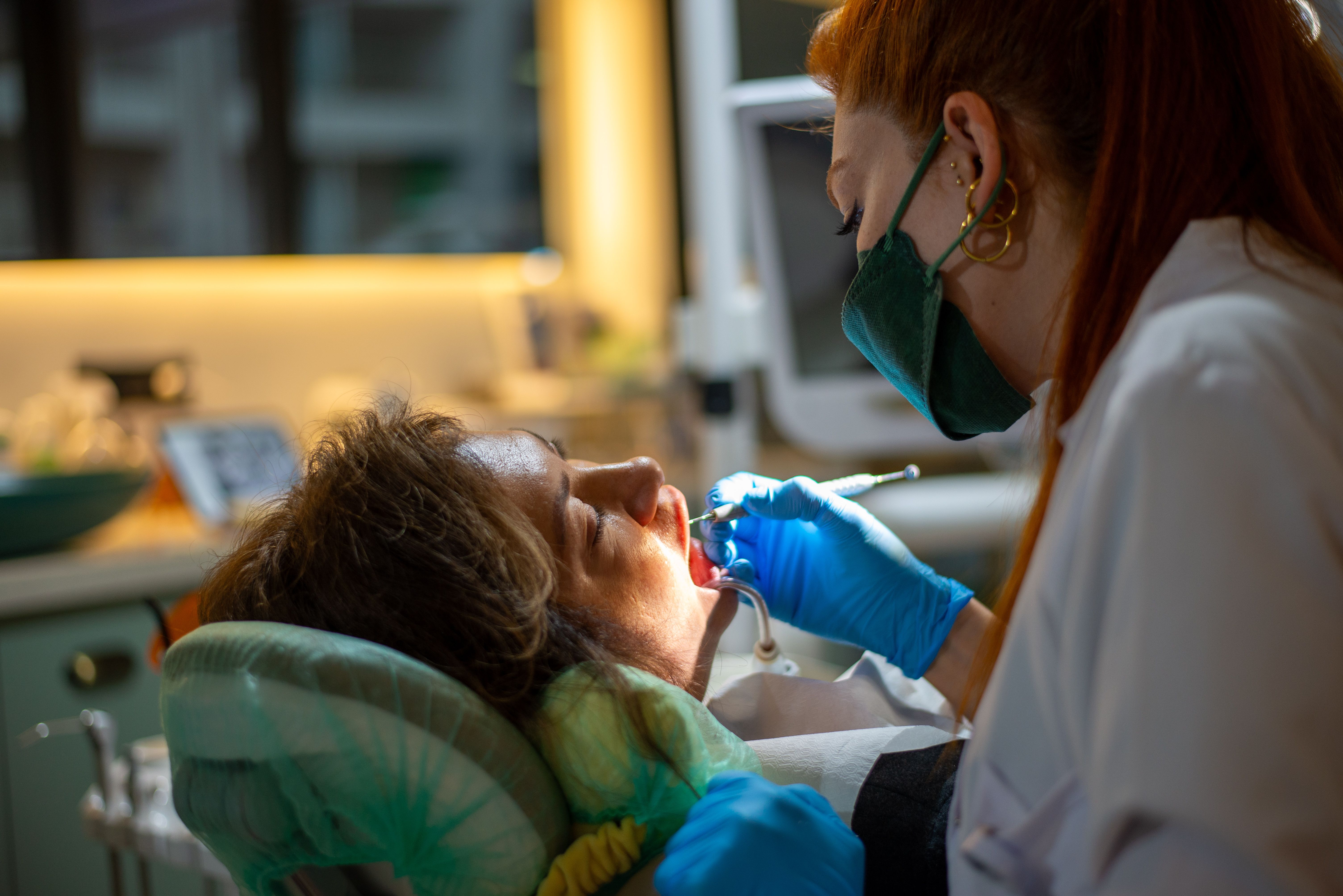 A dentist wearing a mask examines a patient lying down in a dental clinic setting.