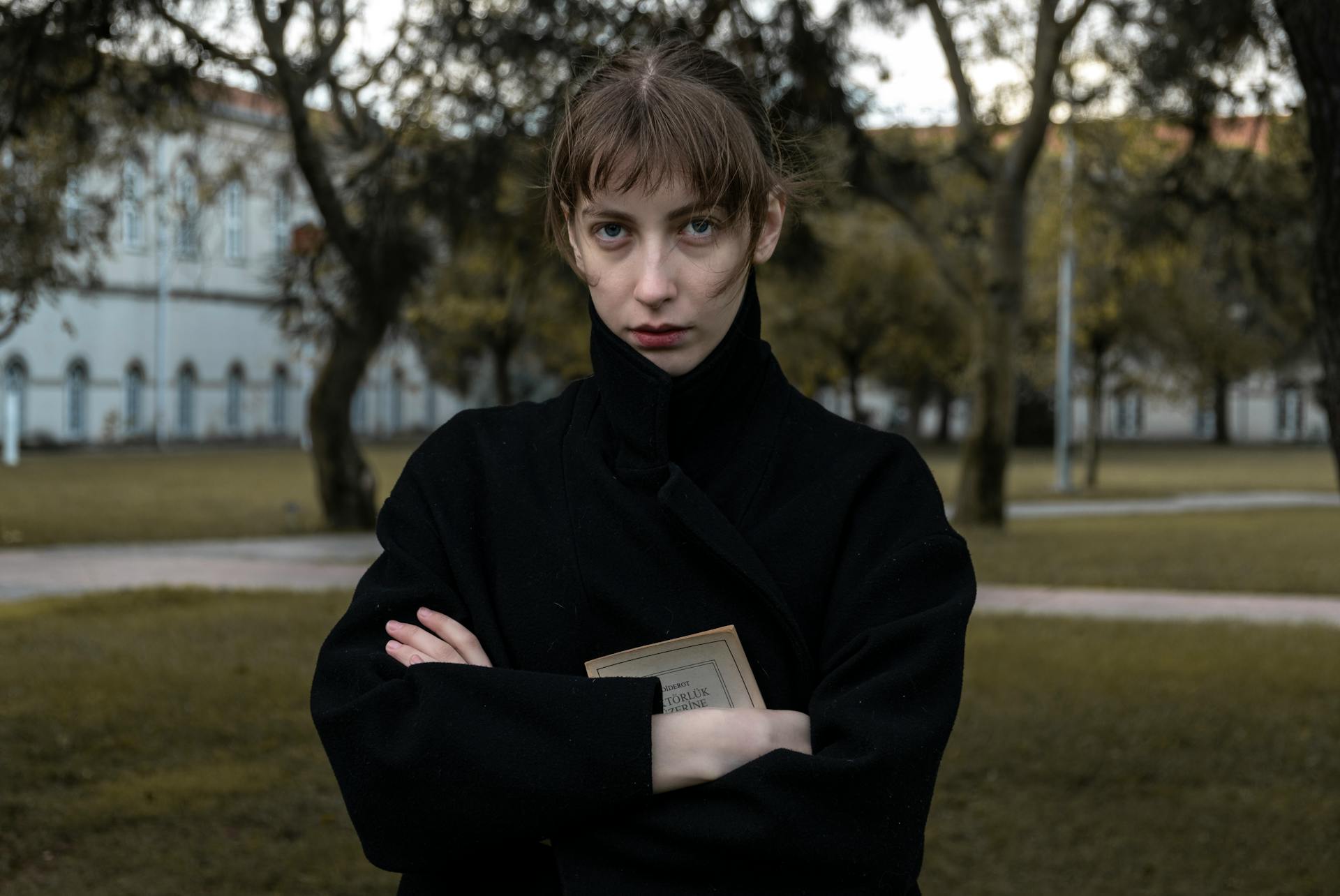 Portrait of a woman in a black coat holding a book in a serene park setting, exuding elegance.