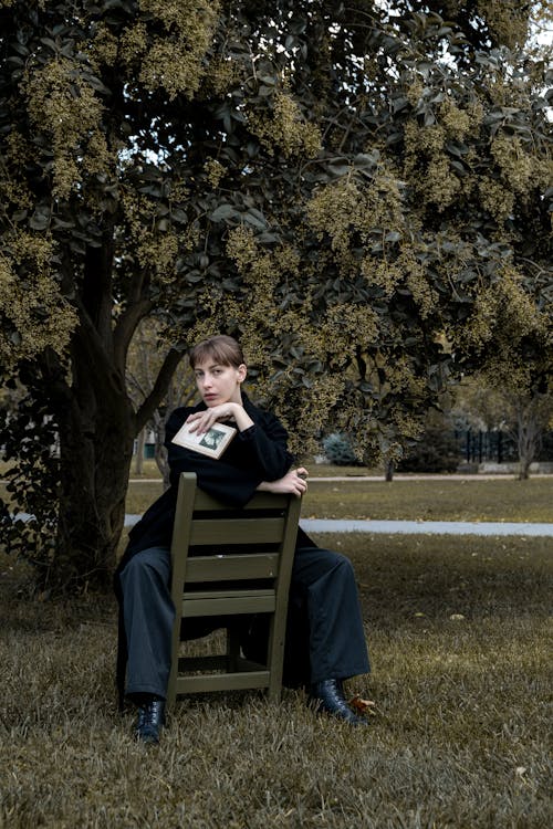 Young Woman in Black Coat and Pants Posing on Chair in Park