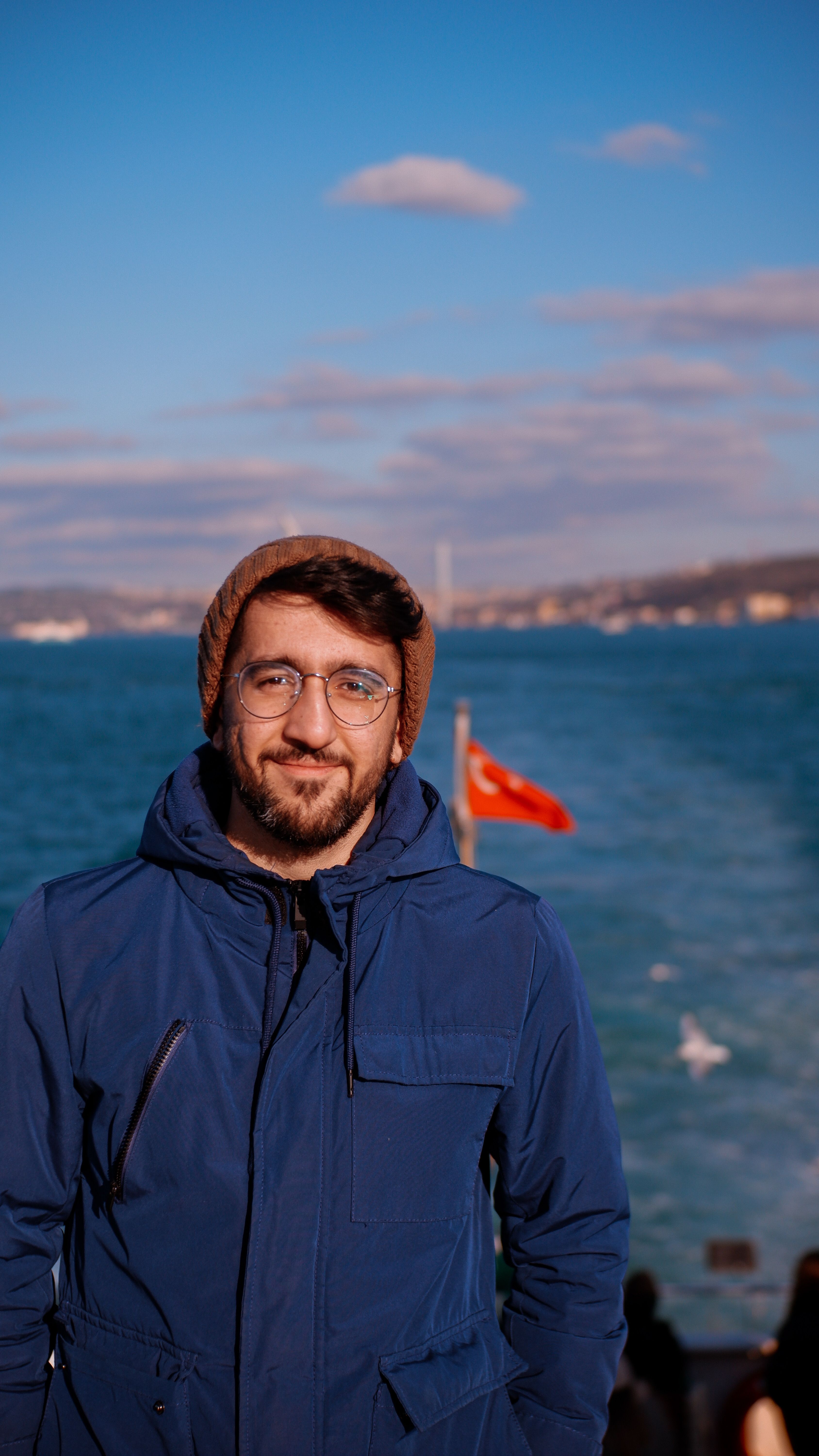 A man in a blue jacket stands by the Bosporus in Istanbul with a view of the sea and Turkish flag.