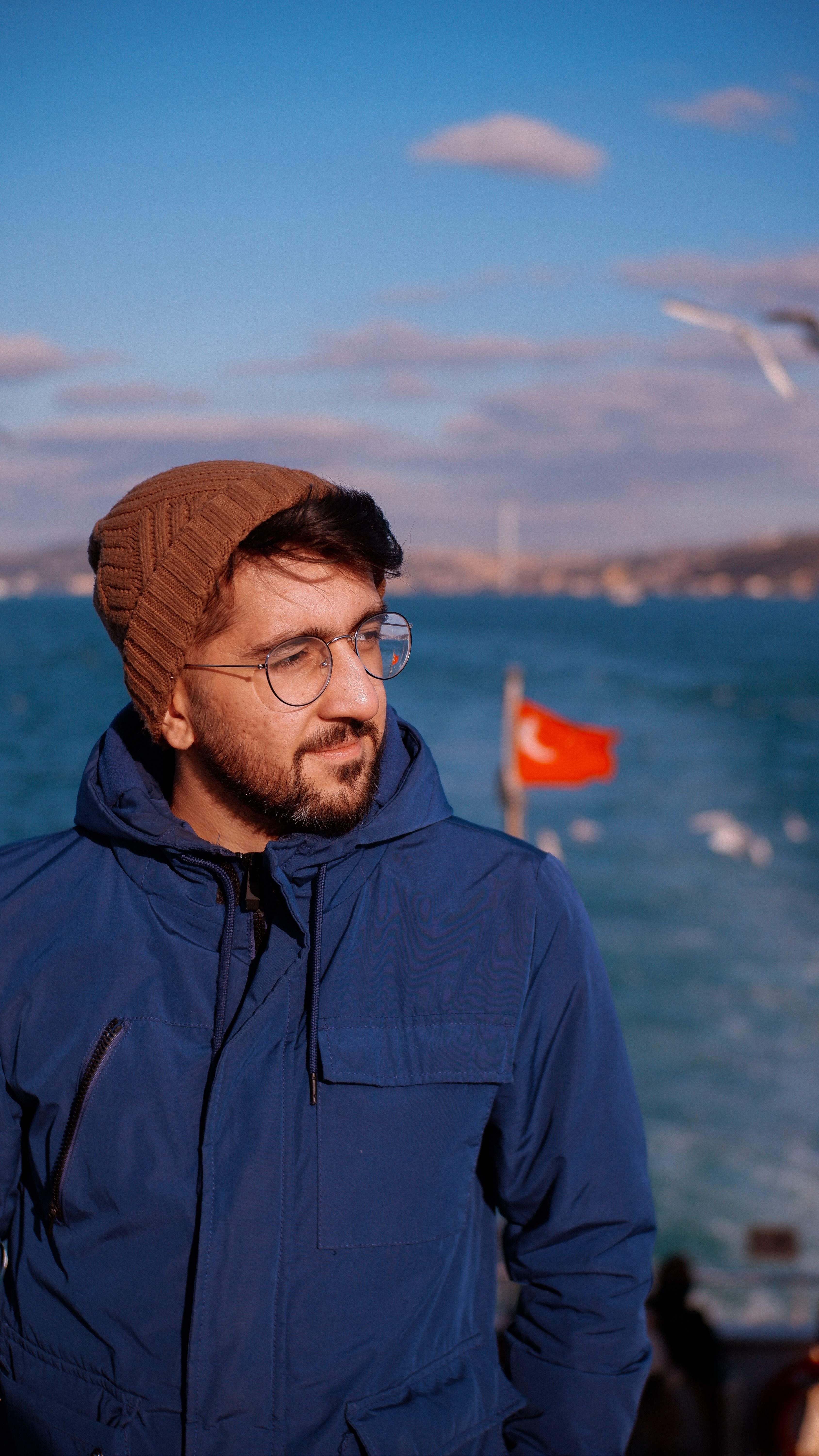 Man in blue jacket and brown hat standing by the Bosporus with a Turkish flag in the background, Istanbul.