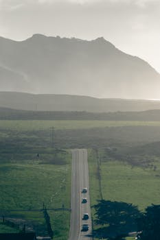 A serene rural road leading through lush fields towards misty mountains under a cloudy sky.