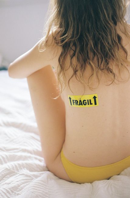 Back view of a woman with a 'Fragile' sticker sitting on a bed in a conceptual photograph.