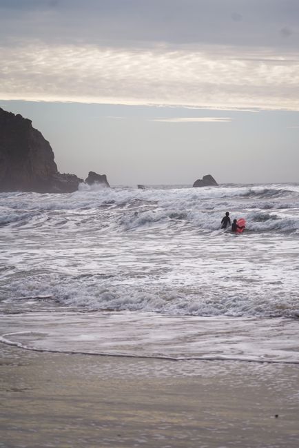 Surfers brave the waves at Cannon Beach, Oregon during a serene sunset with dramatic clouds.