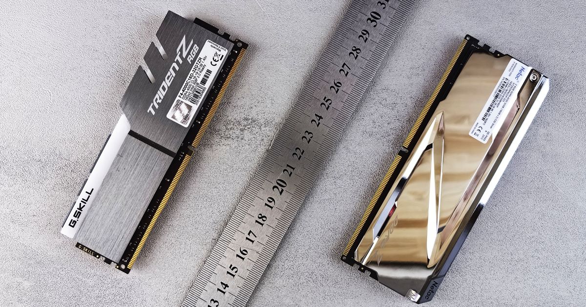 Dois pentes de memória RAM lado a lado sobre uma superfície cinza de concreto, separados por uma régua de aço graduada em centímetros. À esquerda, um G.SKILL Trident Z RGB com dissipador escovado em preto e prata; à direita, um Netac com dissipador espelhado que reflete a luz do ambiente