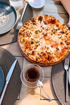 Top view of a freshly baked pizza with a cup of tea served on a rustic wooden table outdoors.