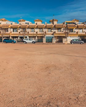 Row of residential townhouses with parked cars and sandy foreground in Santiago de la Ribera, Spain.