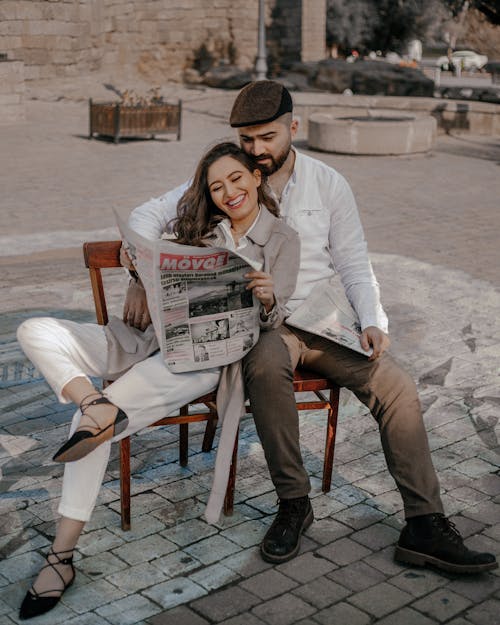 Smiling Couple Sitting on Chairs in City and Reading Newspaper
