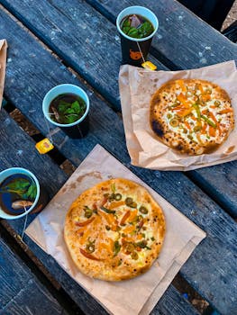 Freshly baked artisan pizzas with vegetables on rustic outdoor table, accompanied by herbal tea.