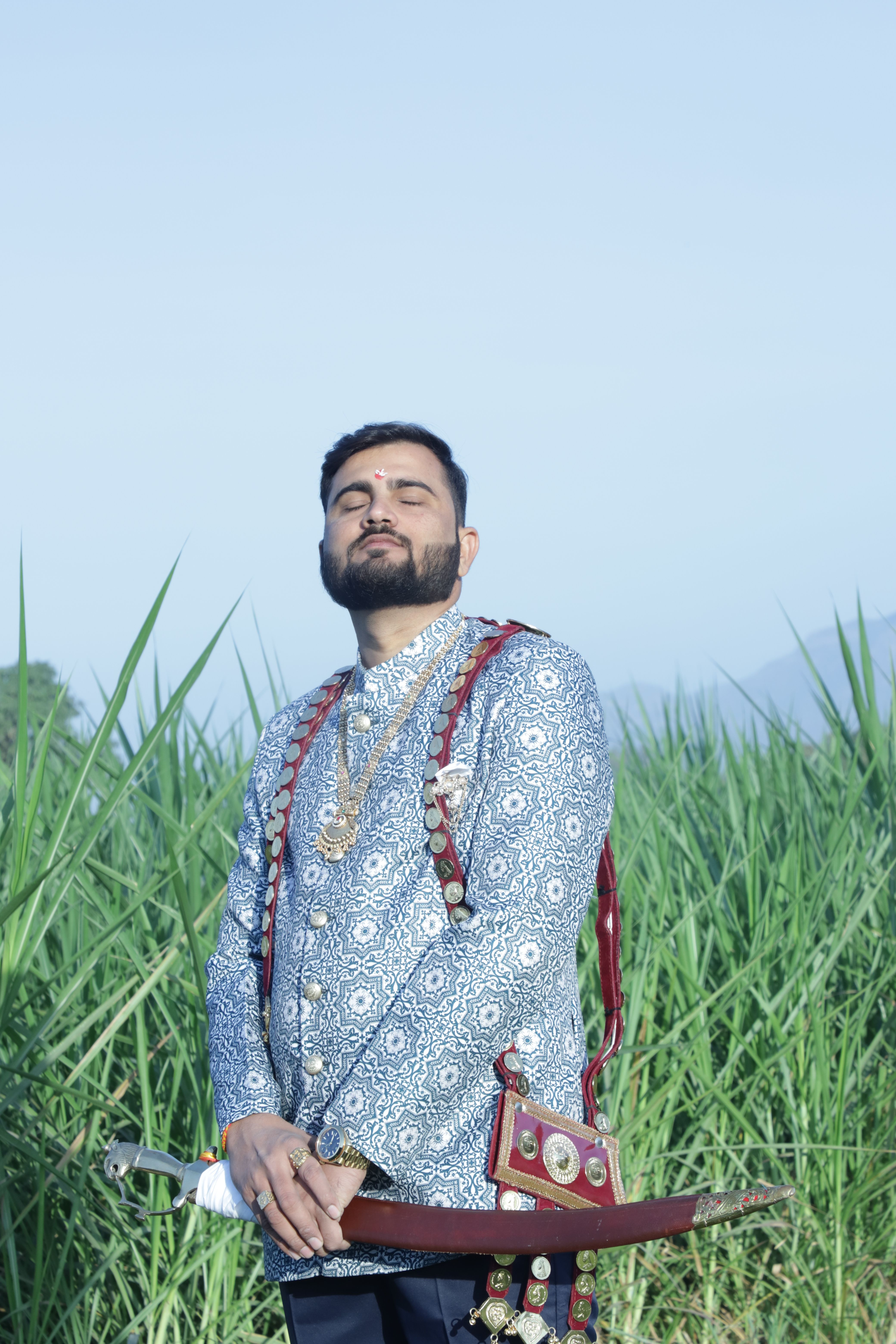 Portrait of an Indian man in traditional clothing holding a sword amidst green fields.