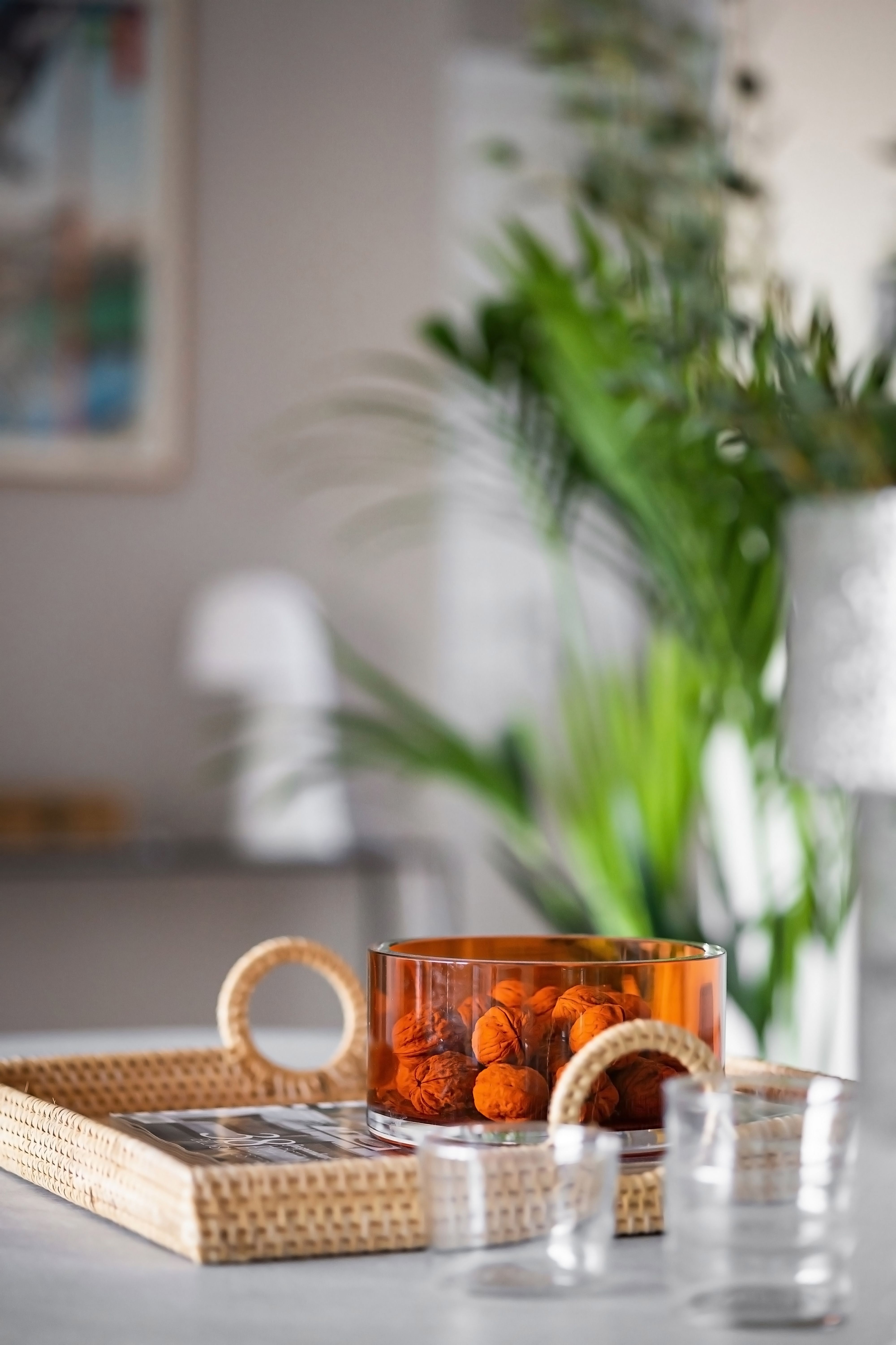 An orange bowl filled with decor sits on a wicker tray against a backdrop of lush greenery indoors.