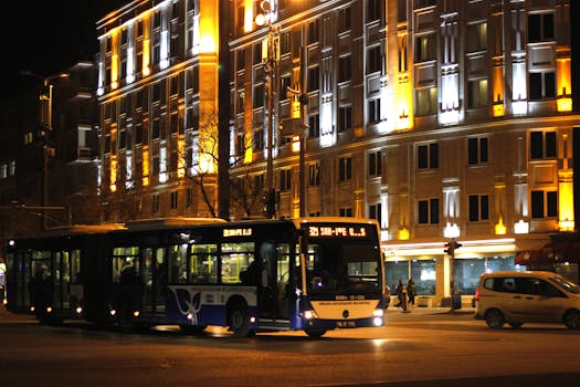 A city street scene captured at night with a brightly illuminated building and a passing public transit bus.