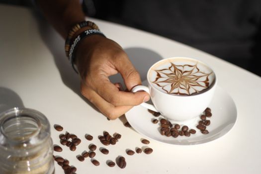 Hand holding a beautifully crafted latte with artistic design, surrounded by scattered coffee beans.