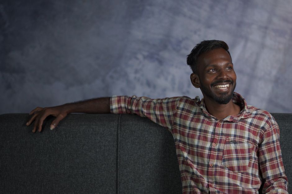 Portrait of a smiling man sitting on a grey couch with a plaid shirt against a textured background.