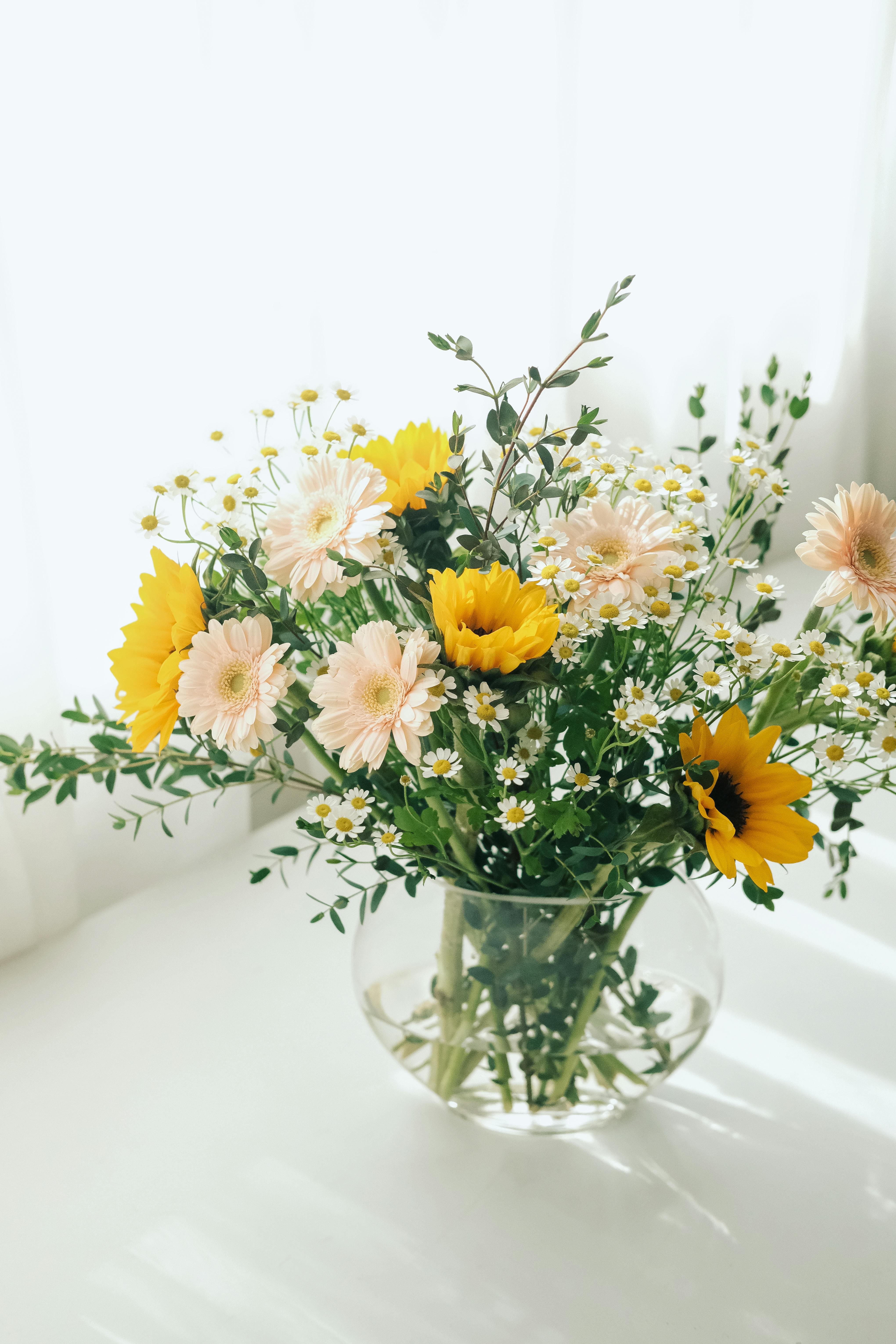 A vibrant bouquet of sunflowers and daisies in a glass vase on a white table.
