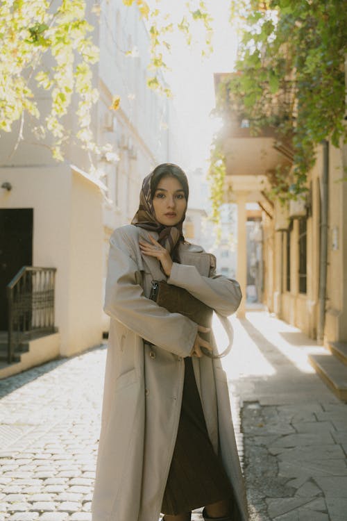 A woman in a beige coat and boots standing on a cobblestone street