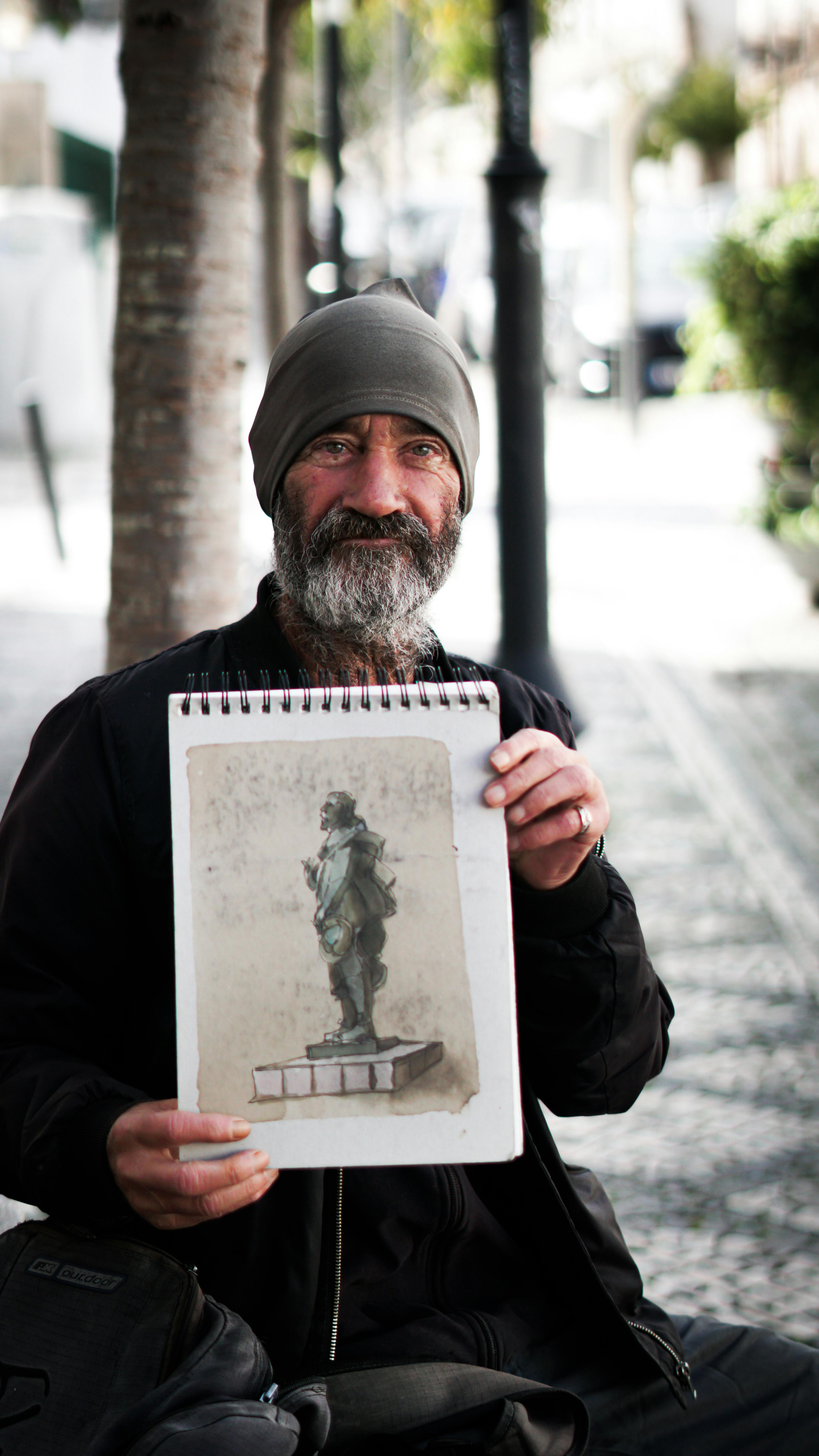 A street artist proudly displays a sketch of a statue while sitting outdoors in Leiria, Portugal.