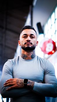 Portrait of a tattooed man in a gray sweatshirt standing indoors with arms crossed.